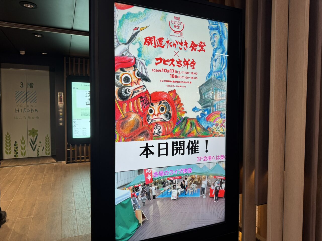 コピス吉祥寺のエレベーターホールにある開運たかさき食堂の電子案内板。だるまのイラストと屋上会場のライブ映像が表示されている