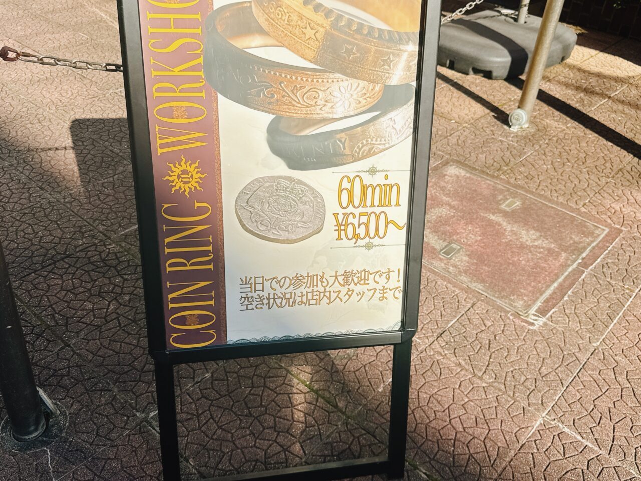 「COIN RING WORKSHOP 60min ¥6,500〜」と書かれたコインリング体験の店頭スタンド看板。精密な模様が刻まれた指輪の写真