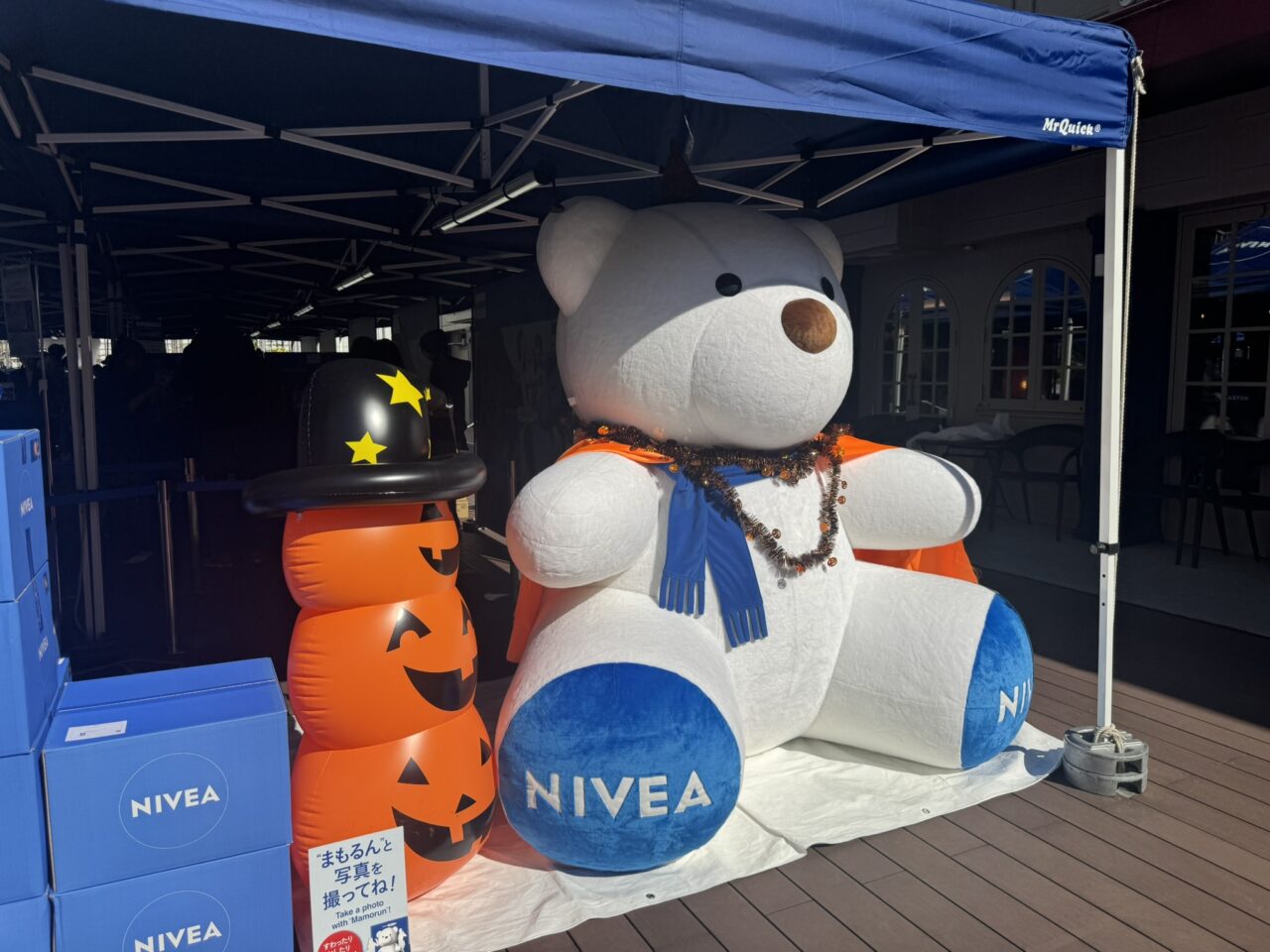 ハロウィンの帽子をかぶったカボチャのバルーンと、オレンジのマントを着た巨大なニベアの白くま「まもるん」